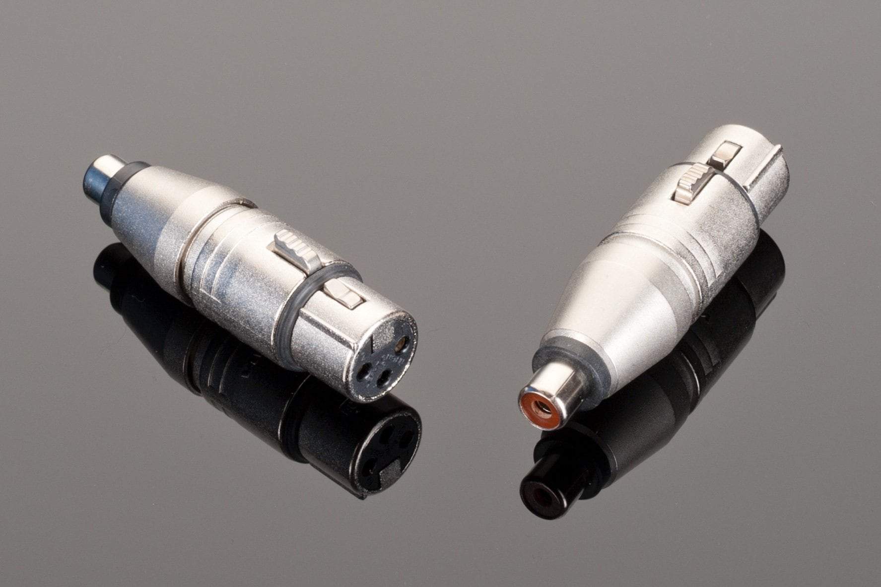 Two Neutrik silver audio connectors, one featuring a female XLR with three pins and another an RCA terminated interconnect with a single pin, are displayed on a reflective black surface.