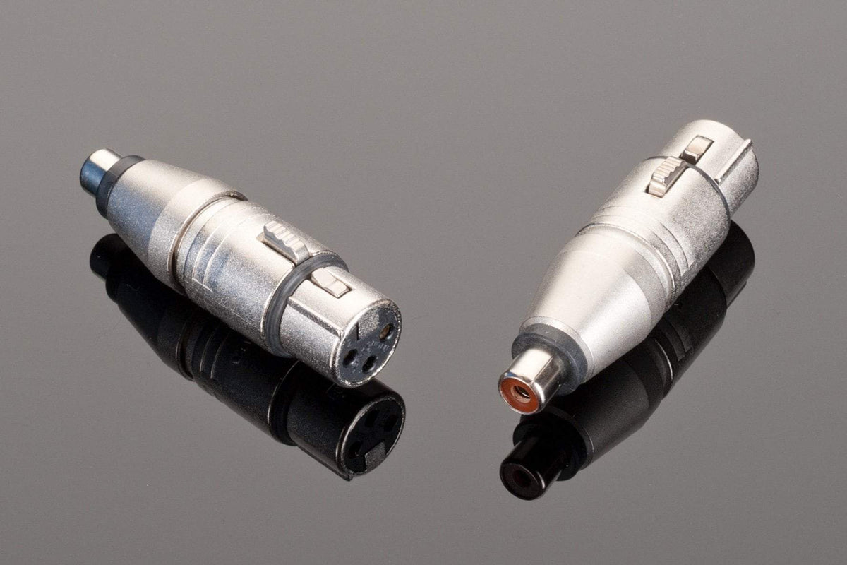 Two Neutrik silver audio connectors, one featuring a female XLR with three pins and another an RCA terminated interconnect with a single pin, are displayed on a reflective black surface.