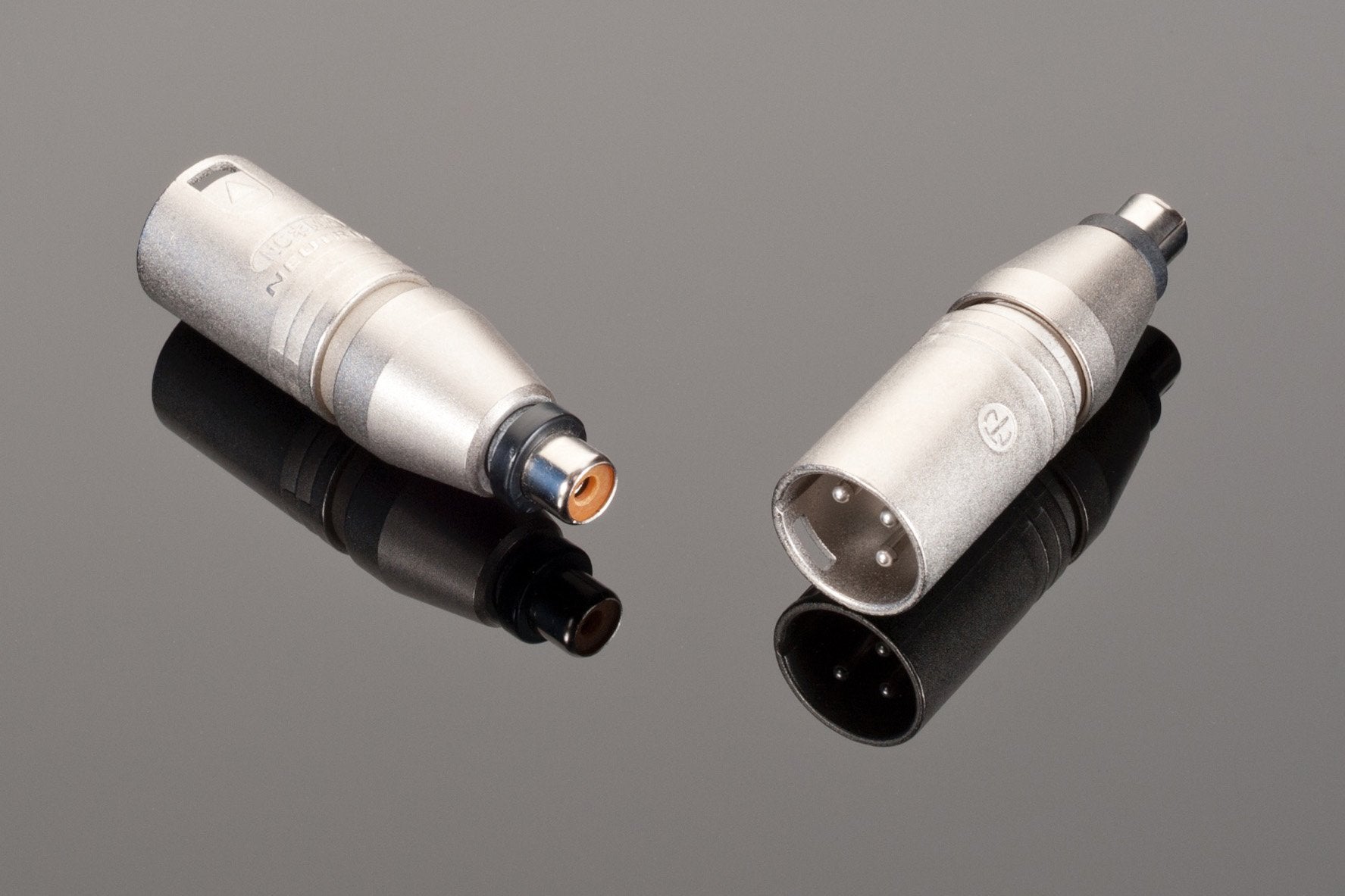Two cylindrical metal connectors, one with a single center pin and the other with three pins, are placed on a reflective surface. The setup almost resembles the sturdy design of the Neutrik Female RCA to Male XLR adapter used in high-quality audio gear.
