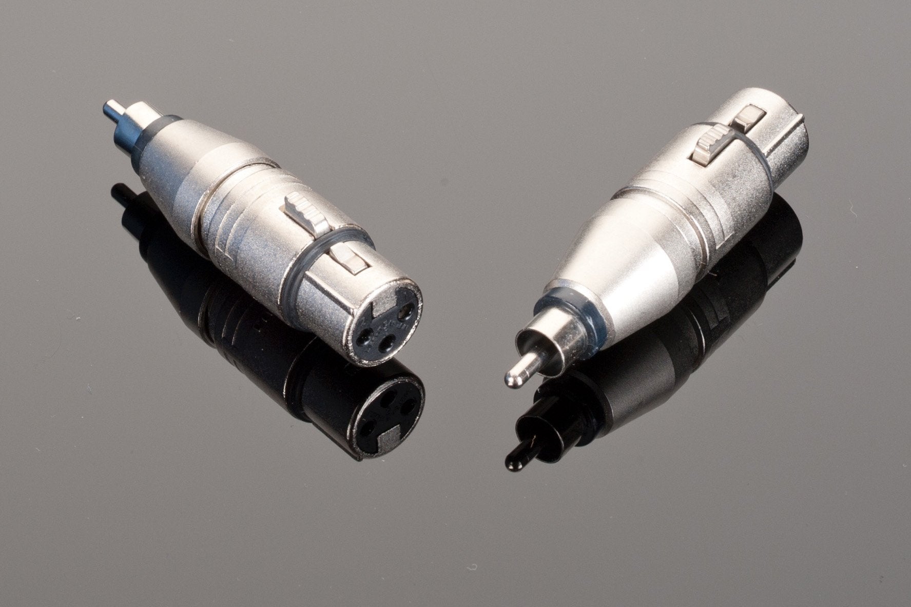Two silver Neutrik Male RCA to Female XLR adapters are placed on a reflective black surface, one displaying the balanced connection XLR end and the other showcasing the RCA terminated interconnect.