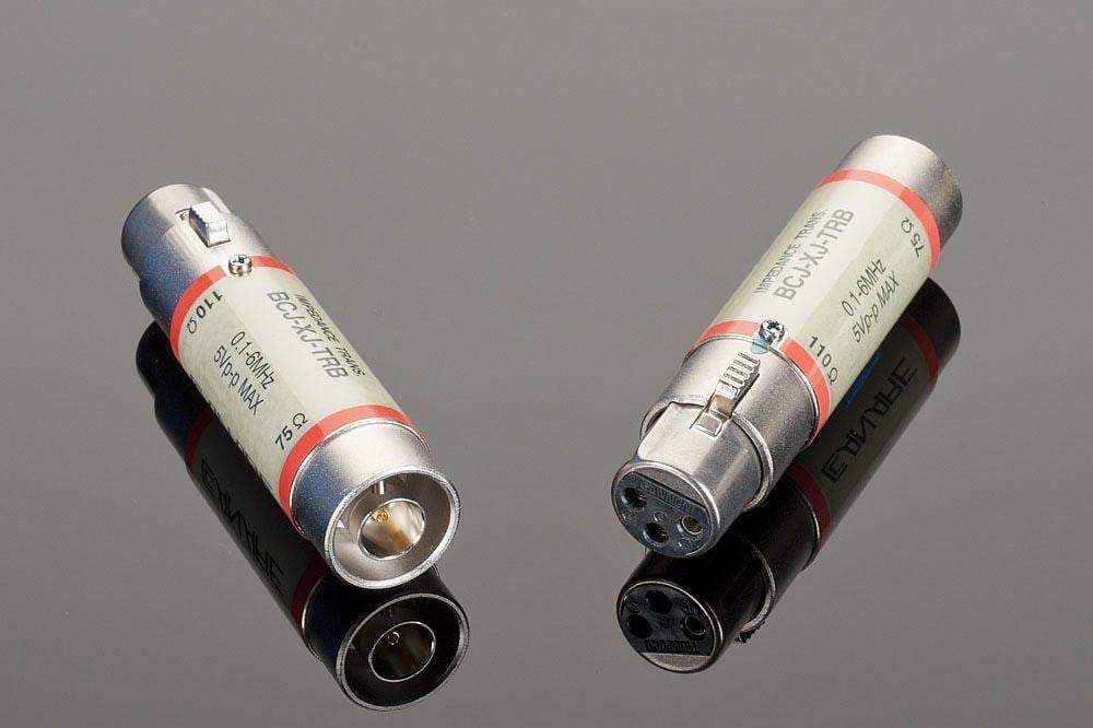Two high-quality Transparent Audio 75-Ohm Female BNC to 110-Ohm Female XLR connectors are laid on a reflective surface, one positioned upright showing its contact pins and the other laying flat displaying its labeled side. Ideal for home entertainment setups requiring reliable digital audio adapters.