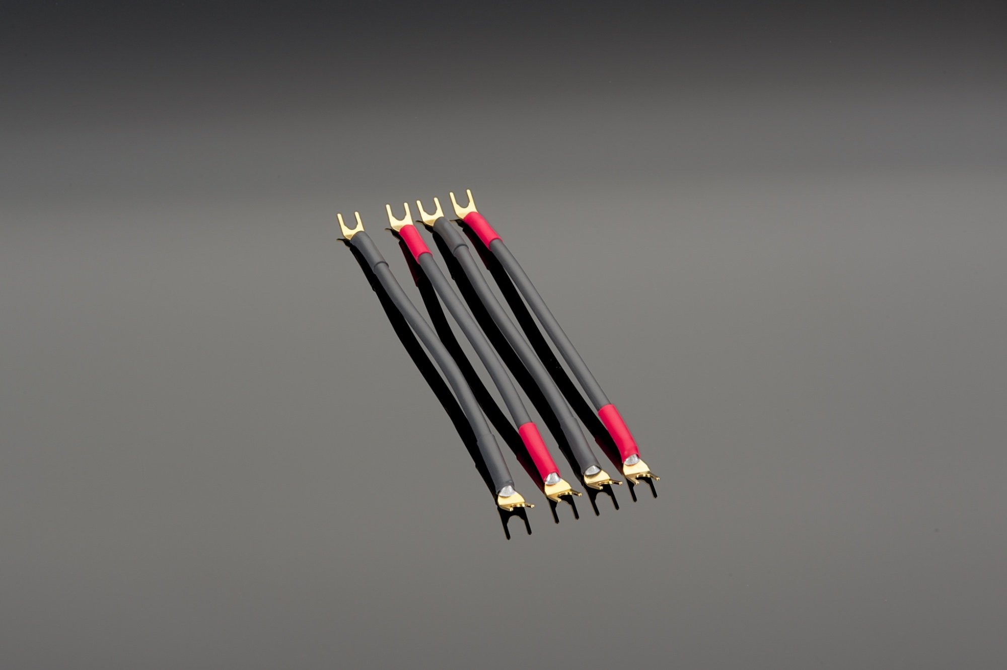 Four Transparent Audio Basic Biwire Adapters, each with black and red connectors featuring forked ends, are arranged in parallel on a gray background. These adapters boast high purity copper strands for superior conductivity.