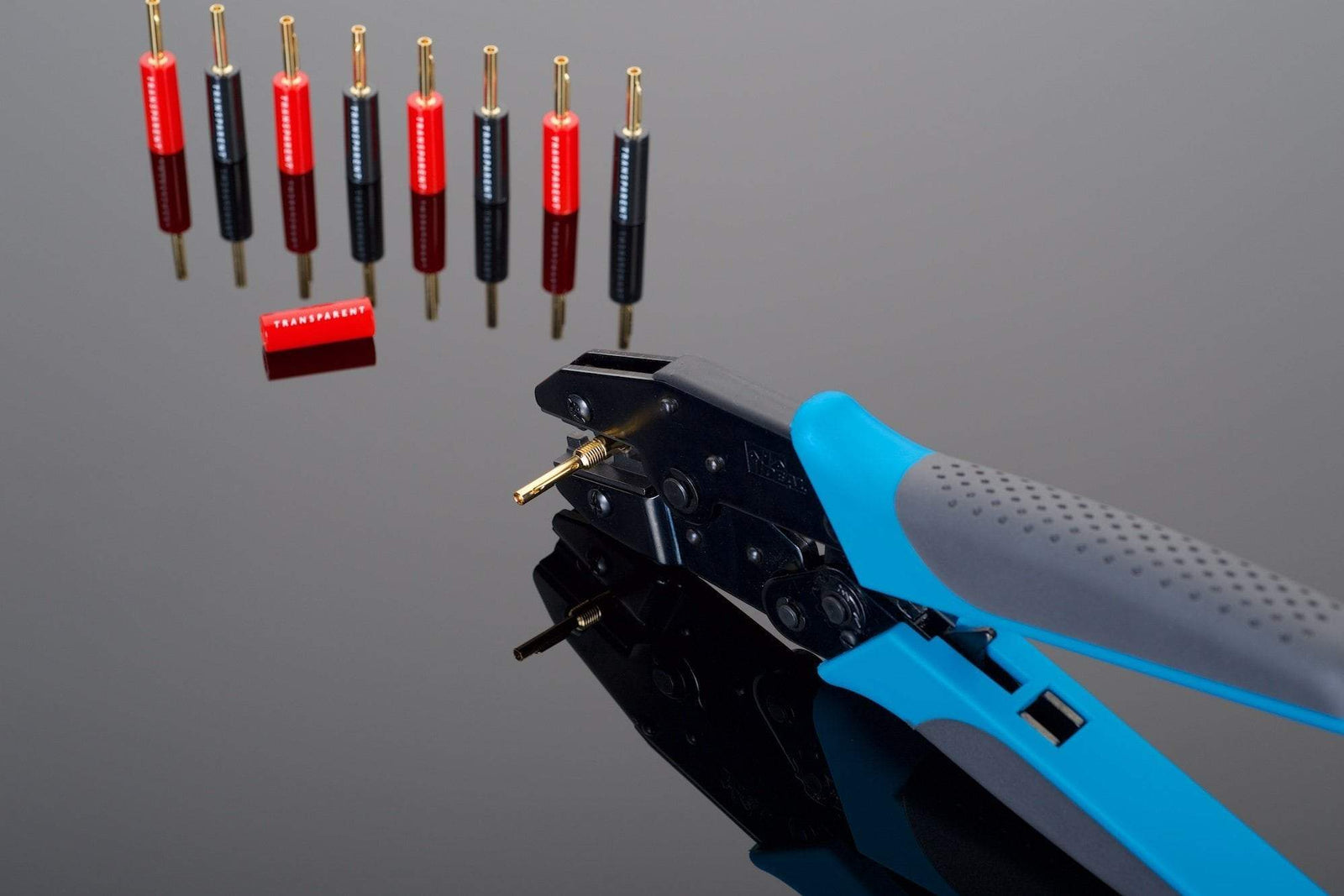 Close-up of the Transparent Audio Crimping Tool, featuring a blue handle with rubber grips, perfectly positioned next to eight banana plugs and one red plastic component on a reflective surface—ideal for prepping your speaker cable.