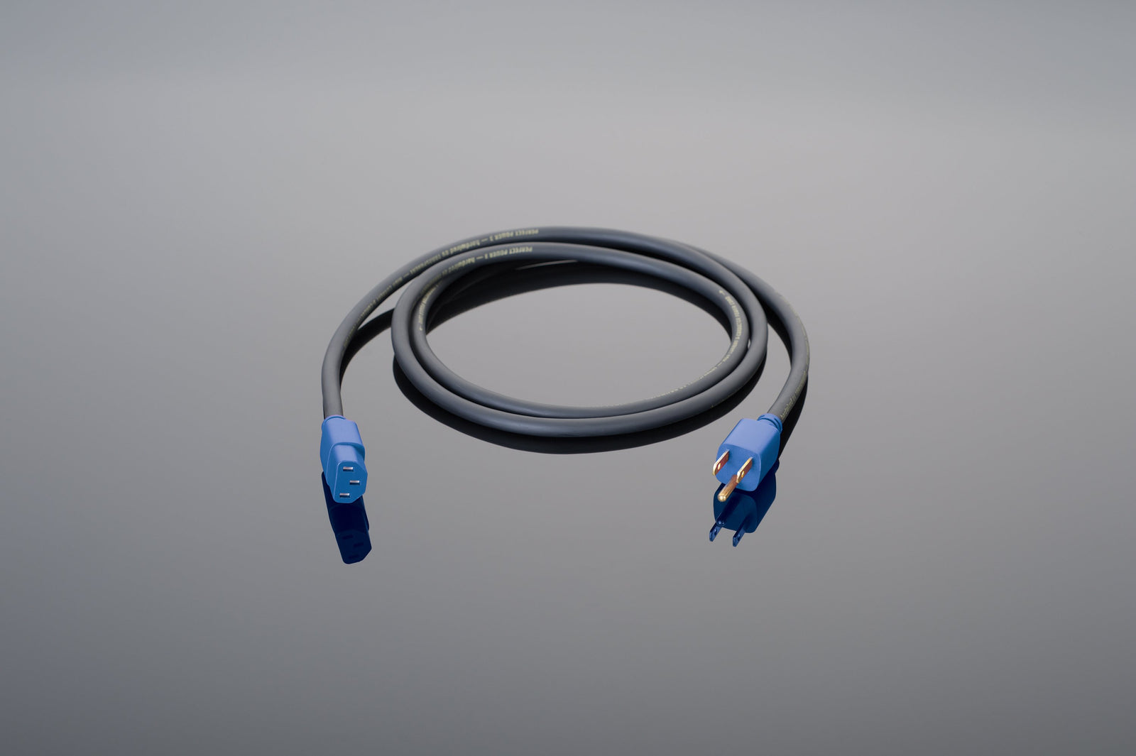 A coiled black Transparent Audio Hardwired 3-Conductor Power Cord with blue connectors on both ends, one featuring prongs and the other designed for an apparatus connection, placed on a reflective surface.