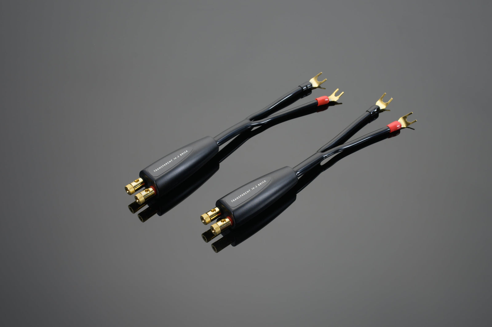 Two Transparent Audio High Performance 14-2 Brick Networks speaker cable adapters with gold-plated connectors and forked terminals, finished in black, are displayed on a gray reflective surface.