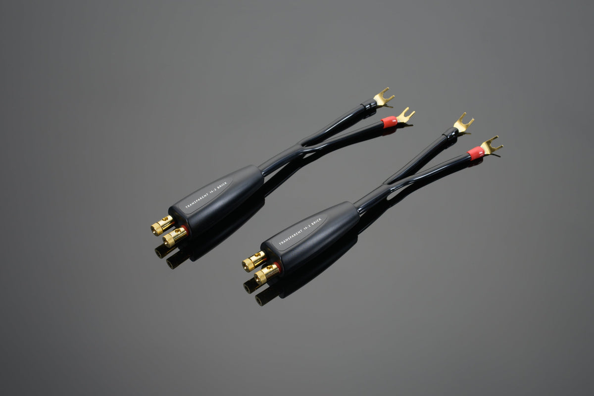 Two black High Performance 14-2 Brick Network speaker cables from Transparent Audio with gold-plated connectors lie parallel to each other on a gray surface. One end has spade connectors, while the other end features banana plugs, making them an essential audio component for any setup.