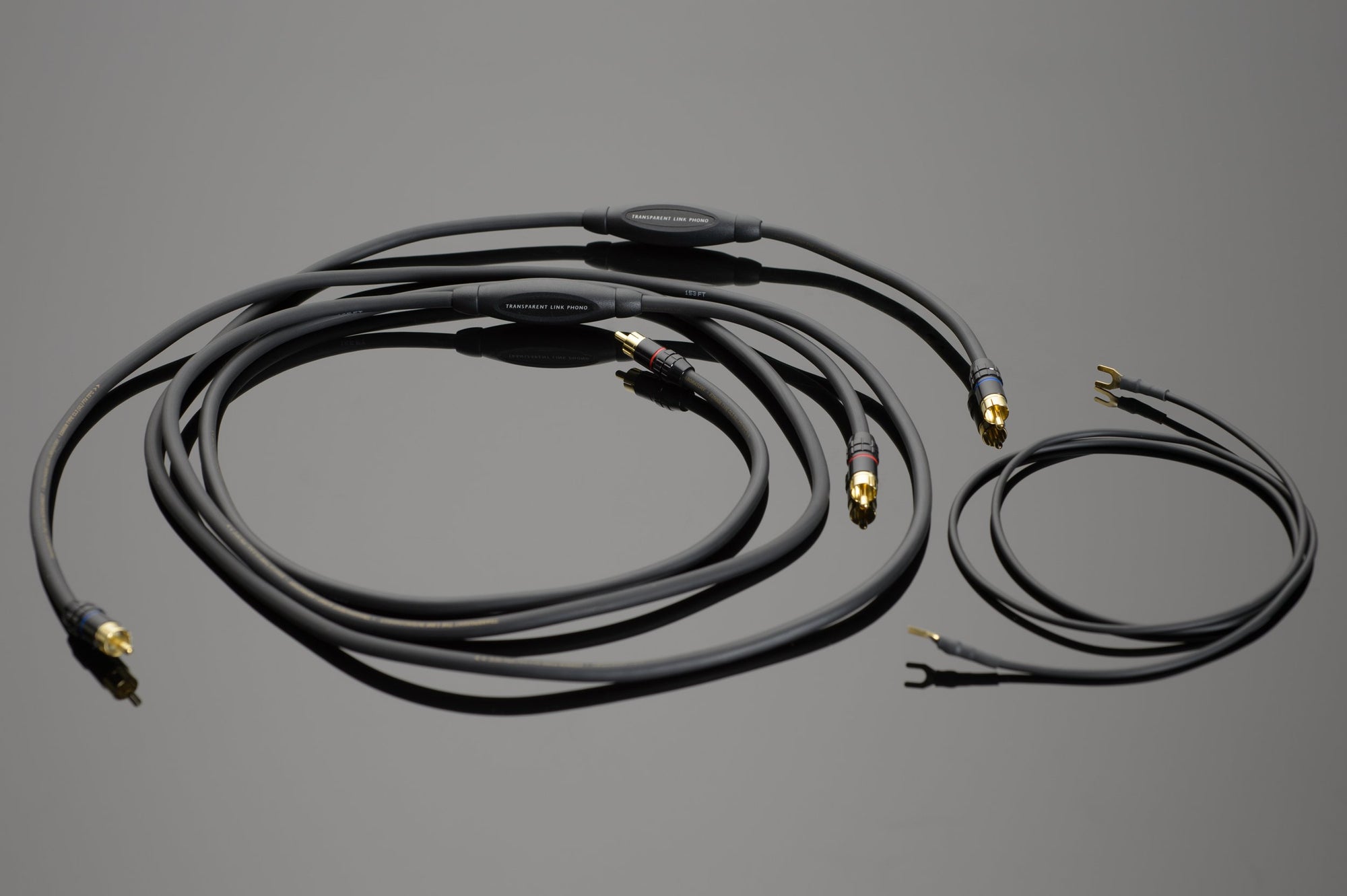 Three Transparent Audio Link Phono Interconnect cables in black, each of different lengths and featuring gold-plated connectors with prong tips, laid out on a grey surface, serve as ideal solutions for a cleaner, clearer sound.