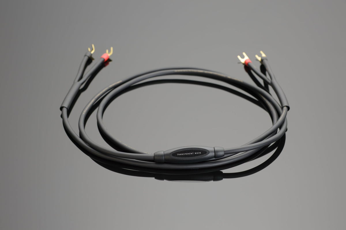 The Wave Speaker Cable by Transparent Audio is a black audio cable featuring four gold-colored connectors, coiled and placed on a grey surface—perfect for any music system.