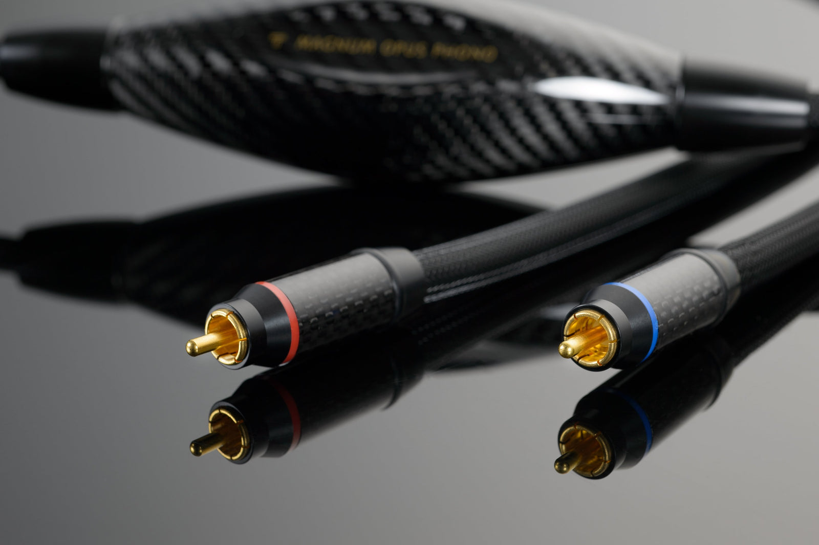Close-up of two black Transparent Audio Magnum OPUS Phono Interconnects with gold-plated connectors, one with a red ring and the other with a blue ring, placed on a reflective black surface.