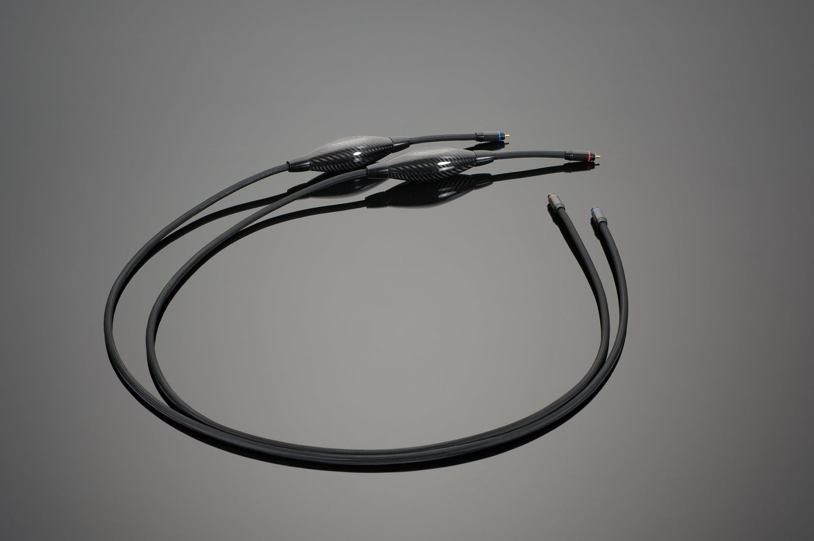 A black, dual-ended audio cable with two connectors on each end, featuring cylindrical protective casings in the middle, is placed on a flat, reflective surface. This exceptional Magnum OPUS Phono Interconnect from Transparent Audio seamlessly pairs with your phono cartridges for pristine sound quality.