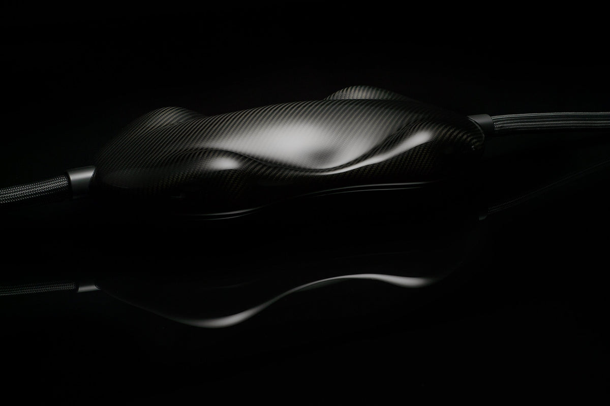 A glossy, black carbon fiber object with a smooth, contoured surface connected to Transparent Audio MAGNUM OPUS Speaker Cable is set against a dark background.