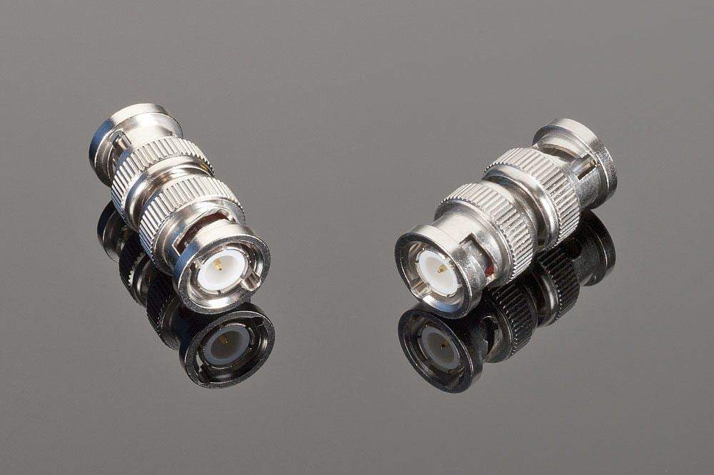 Two high-quality Transparent Audio Male BNC to Male BNC connectors are placed against a reflective surface. The connectors, often used as digital audio adapters in home entertainment setups, are cylindrical with knurled sides.