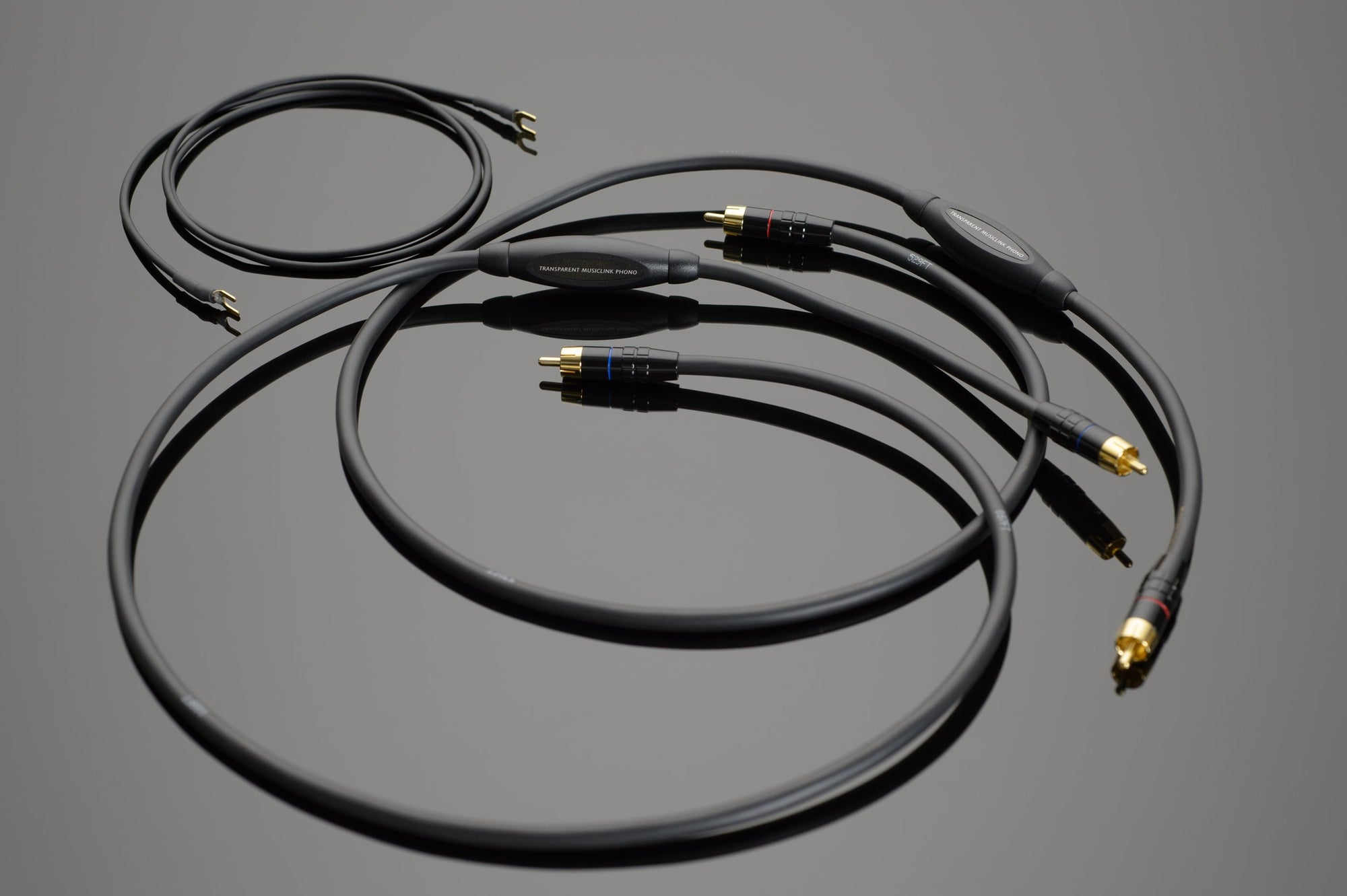 Three black Transparent Audio MusicLink Phono Interconnects with gold-plated connectors are coiled on a reflective gray surface. The cables, ideal for your phonograph set-up, feature labels with text.
