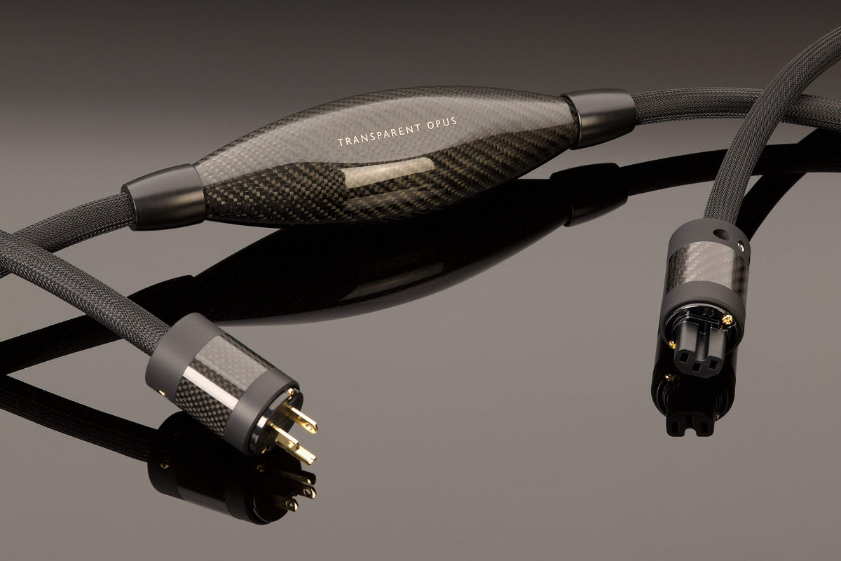 A high-end audio power cable with a carbon fiber casing and gold-plated connectors, labeled the &quot;OPUS Power Cord&quot; by Transparent Audio, is displayed against a reflective surface. Designed for high-resolution amplifiers, it ensures a noise-free power connection for pristine sound quality.