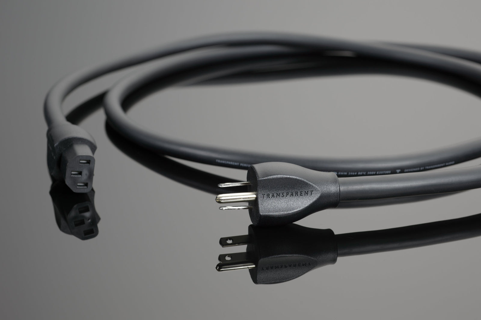The Transparent Audio Performance Power Cord, featuring a coiled black design with a three-prong plug and connector, placed on a reflective surface, offers an upgraded basic design for low-noise power connections, making it ideal for enhanced music and film systems.