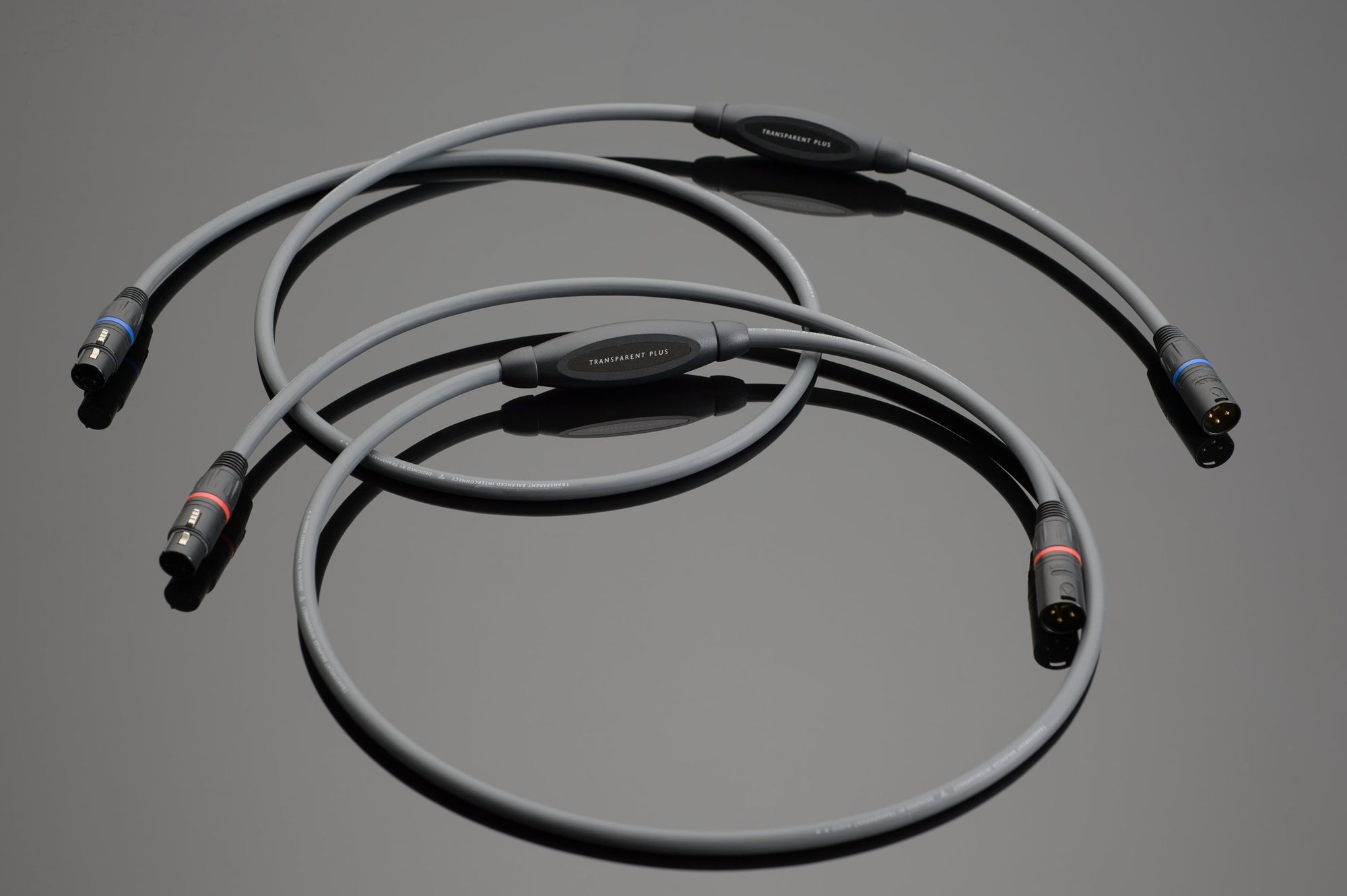 Two Transparent Audio PLUS Balanced Interconnects with black connectors, featuring blue and red color bands near the ends, are placed on a reflective surface.