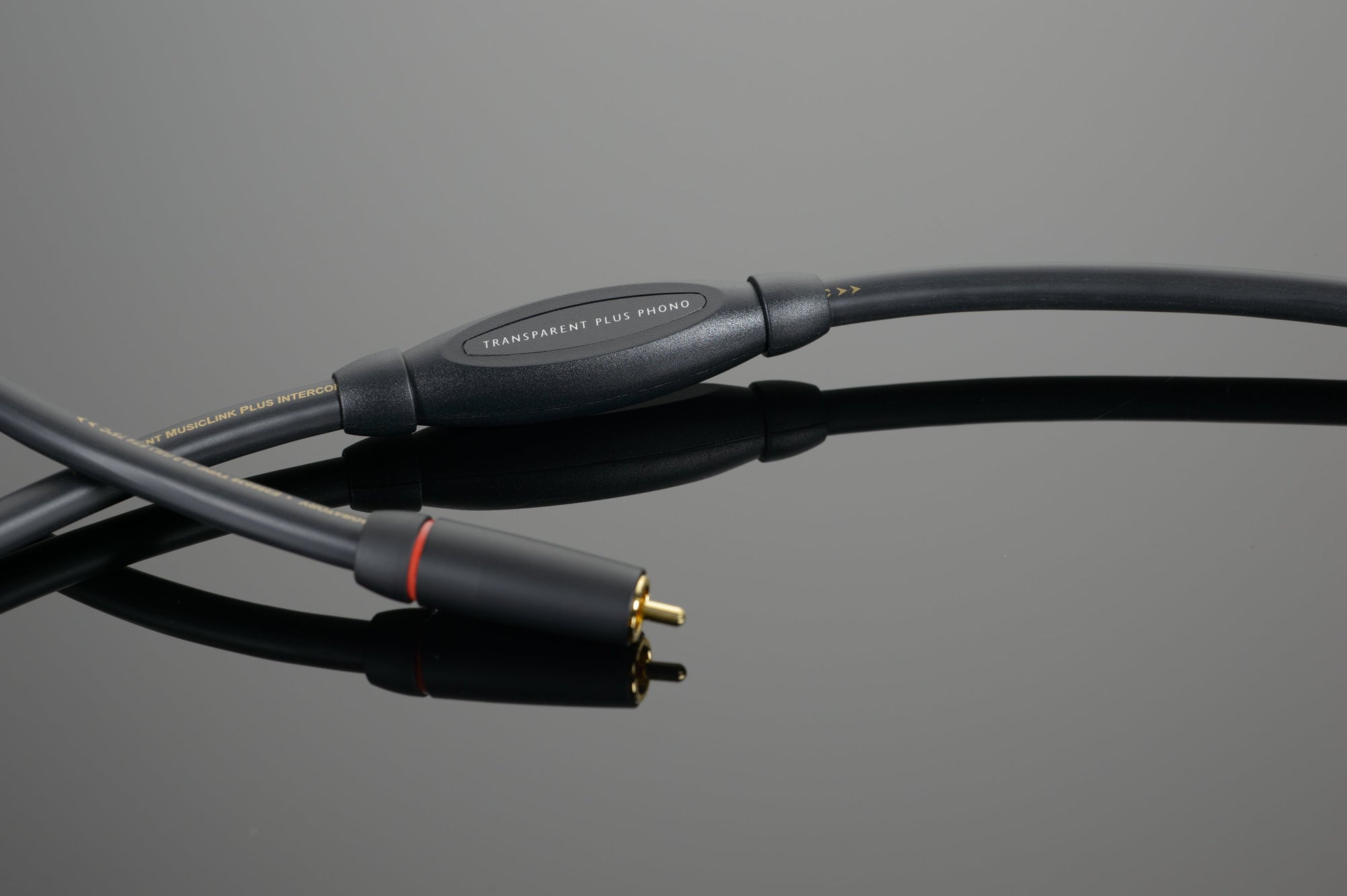 Close-up of a black audio cable with a red ring near one of the connectors, on a reflective gray surface. The essential cable for any phonograph set-up is labeled "Transparent Audio PLUS Phono Interconnect.