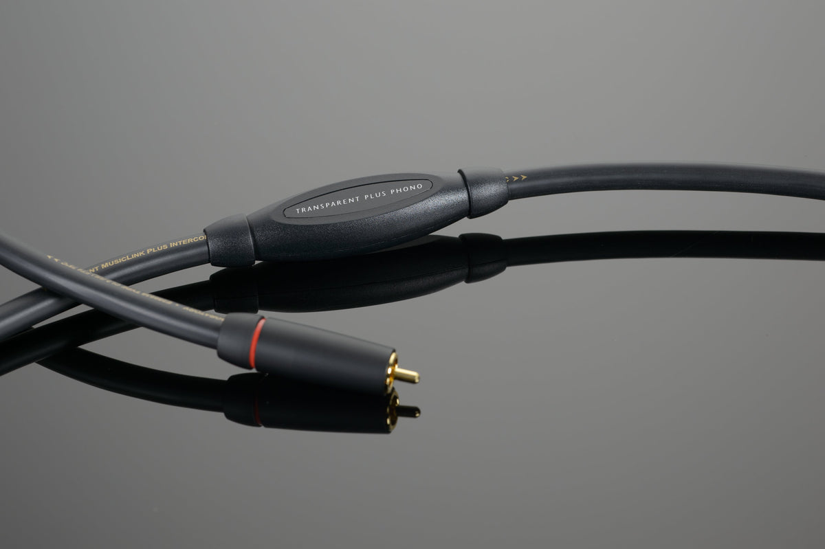 Close-up of a black audio cable with a red ring near one of the connectors, on a reflective gray surface. The essential cable for any phonograph set-up is labeled &quot;Transparent Audio PLUS Phono Interconnect.