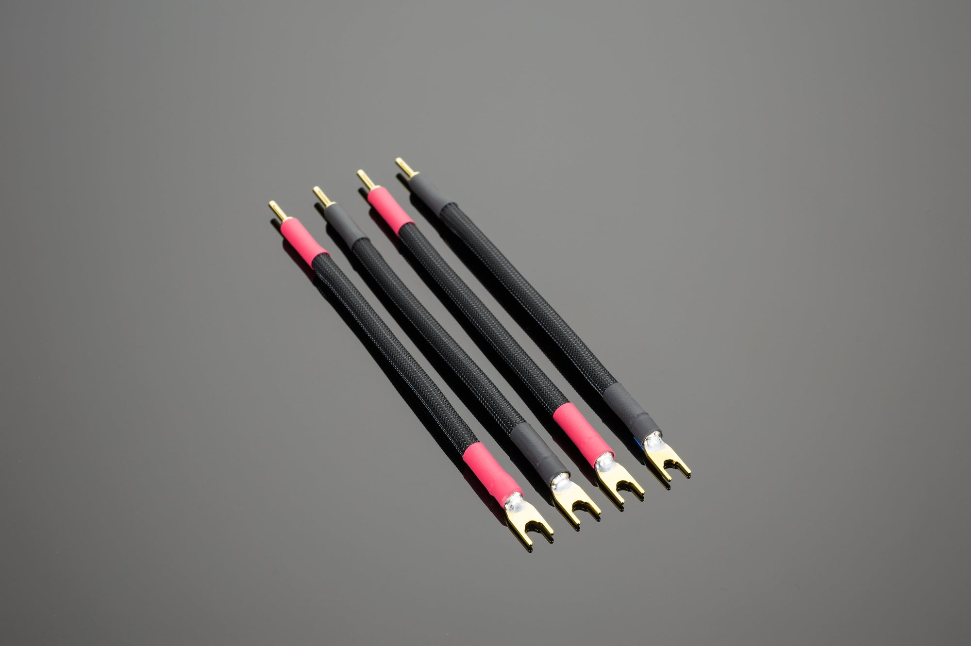 Four Transparent Audio Premium Biwire Speaker Adapters with gold connectors are arranged parallel on a gray surface for high performance connectivity, featuring black and red spade or banana speaker cables.