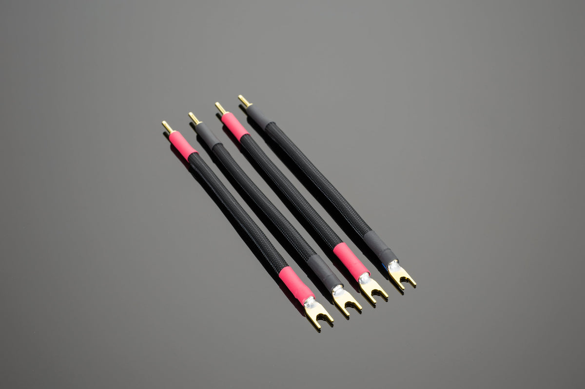 Four Transparent Audio Premium Biwire Speaker Adapters with gold connectors are arranged parallel on a gray surface for high performance connectivity, featuring black and red spade or banana speaker cables.