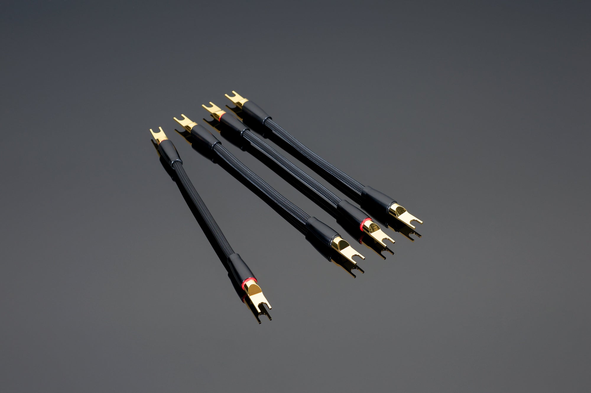 Five Transparent Audio Reference or XL Biwire Adapters, in black and gold with forked connectors and high purity copper strands, arranged diagonally on a dark gray background.