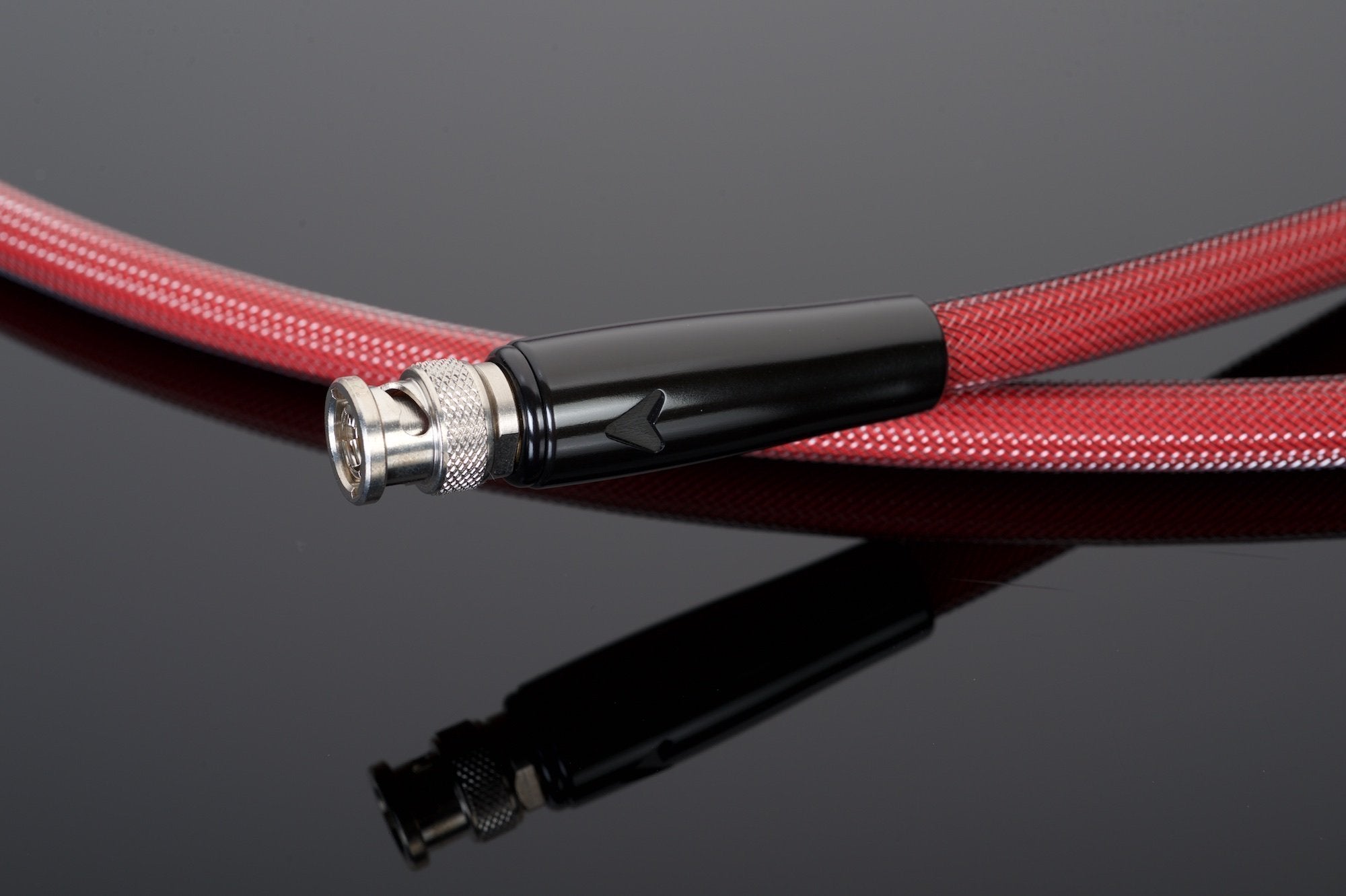 A red braided coaxial cable with a silver BNC connector end, handcrafted in the USA by Transparent Audio, lies coiled on a reflective black surface. Renowned for its superior 75-ohm impedance stability, this REFERENCE 75-Ohm Digital Link cable offers top-tier performance.