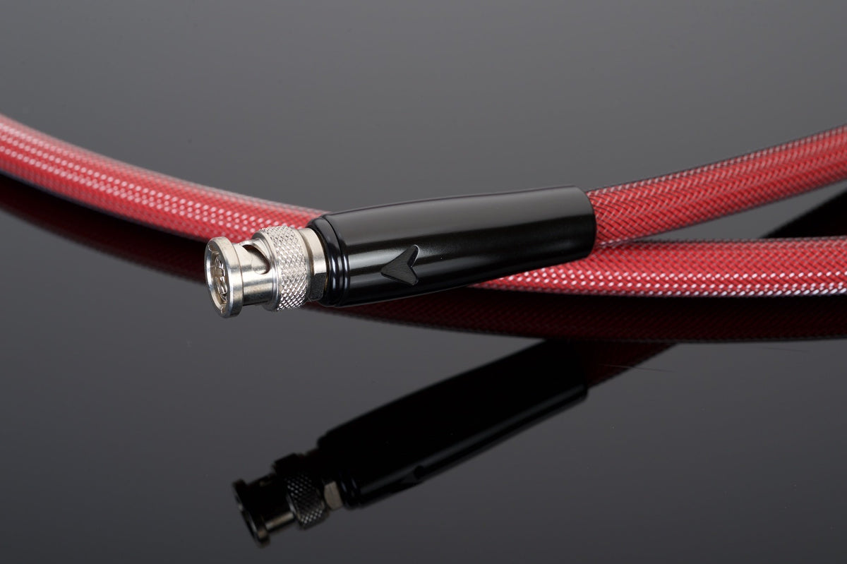 A red braided coaxial cable with a silver BNC connector end, handcrafted in the USA by Transparent Audio, lies coiled on a reflective black surface. Renowned for its superior 75-ohm impedance stability, this REFERENCE 75-Ohm Digital Link cable offers top-tier performance.
