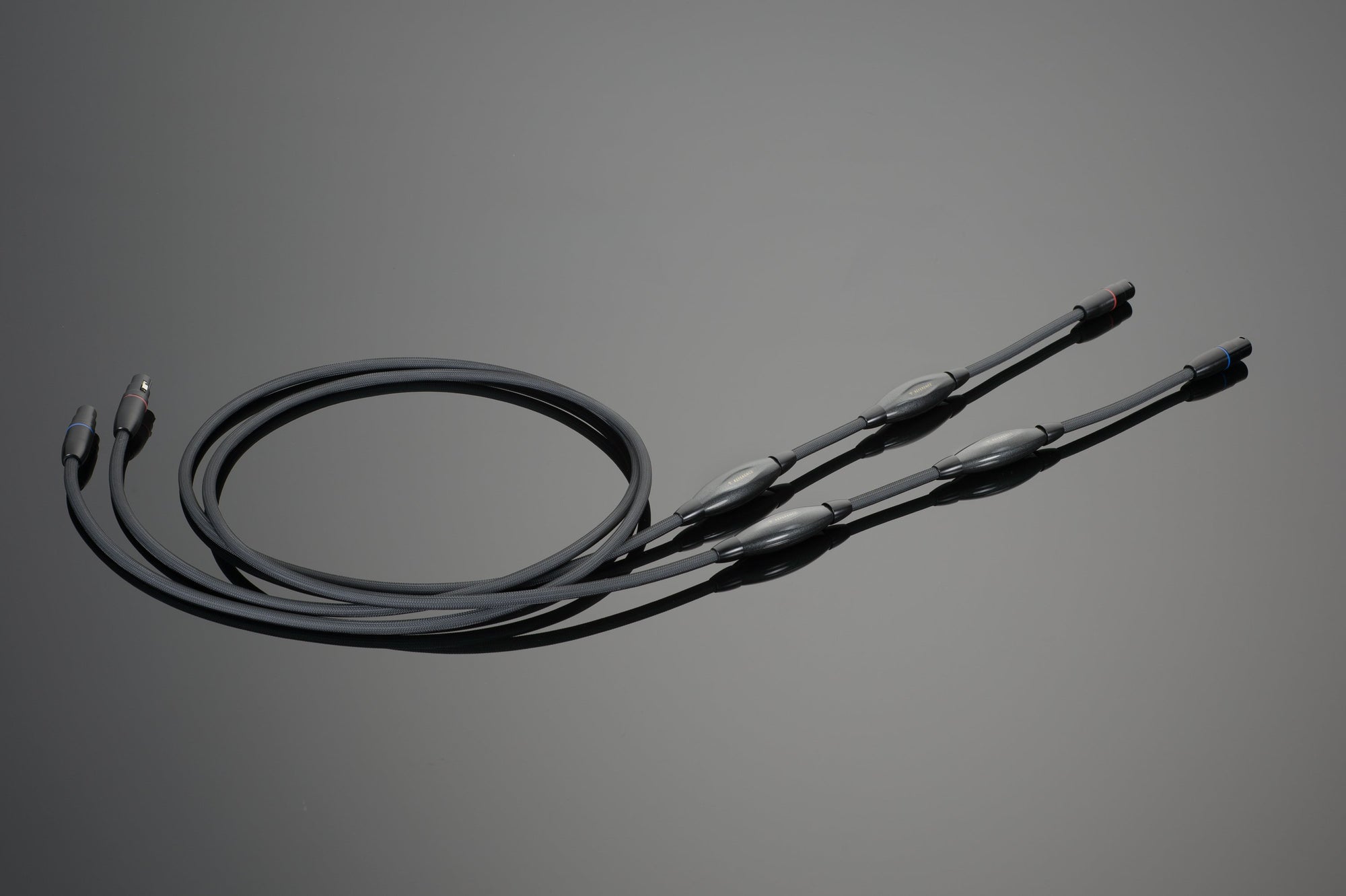The REFERENCE Balanced Interconnect set by Transparent Audio, featuring black audio cables with connectors at each end and cylindrical bumps along their length, is coiled neatly on a grey surface—an ideal choice for audiophiles seeking high-resolution sound.