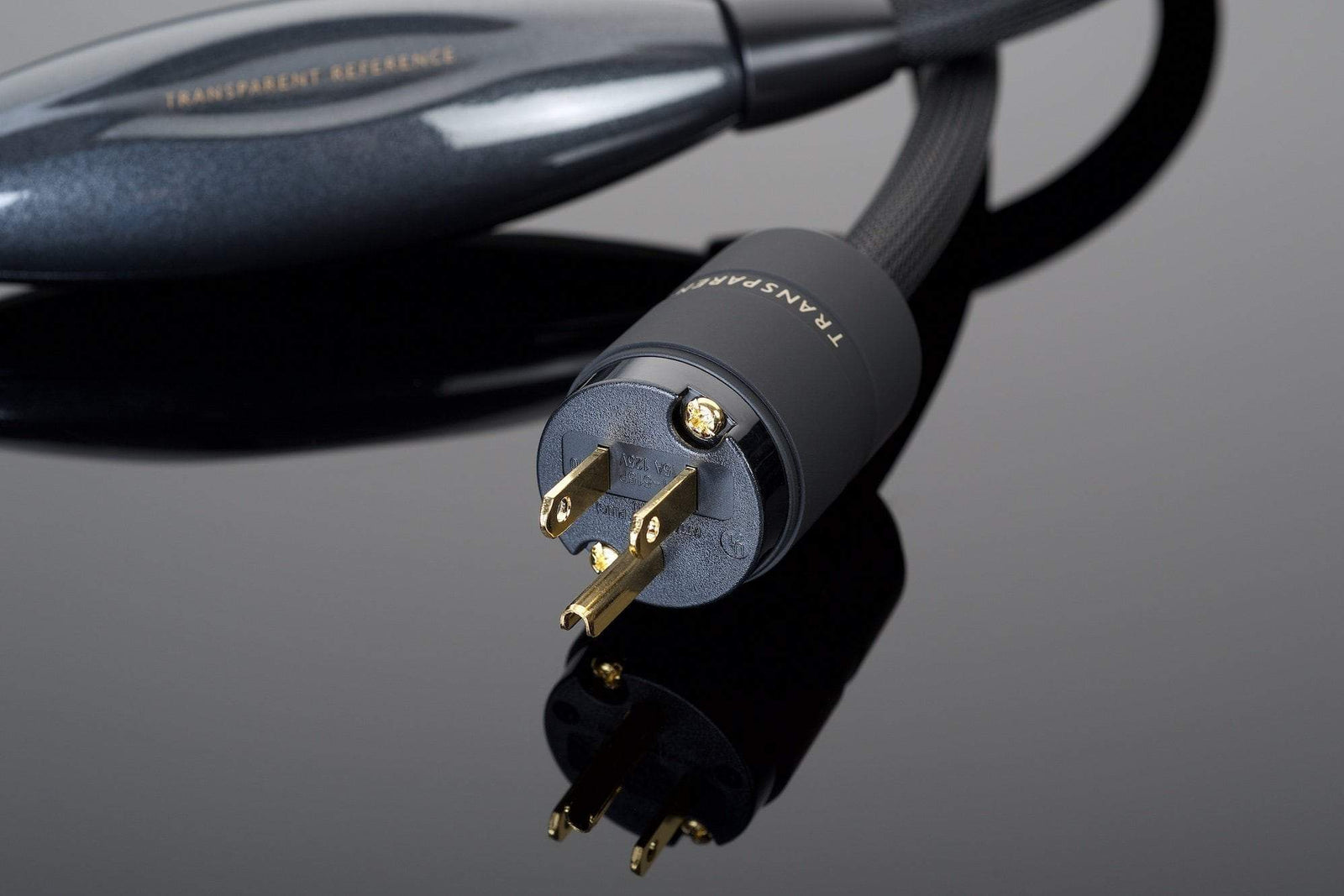 Close-up of a high-end OFHC power cable connector placed on a glossy black surface. The REFERENCE Power Cord by Transparent Audio features gold-plated prongs and is labeled with "TRANSPARENT" and "REFERENCE," ensuring a noise-free power connection for your premium audio setup.