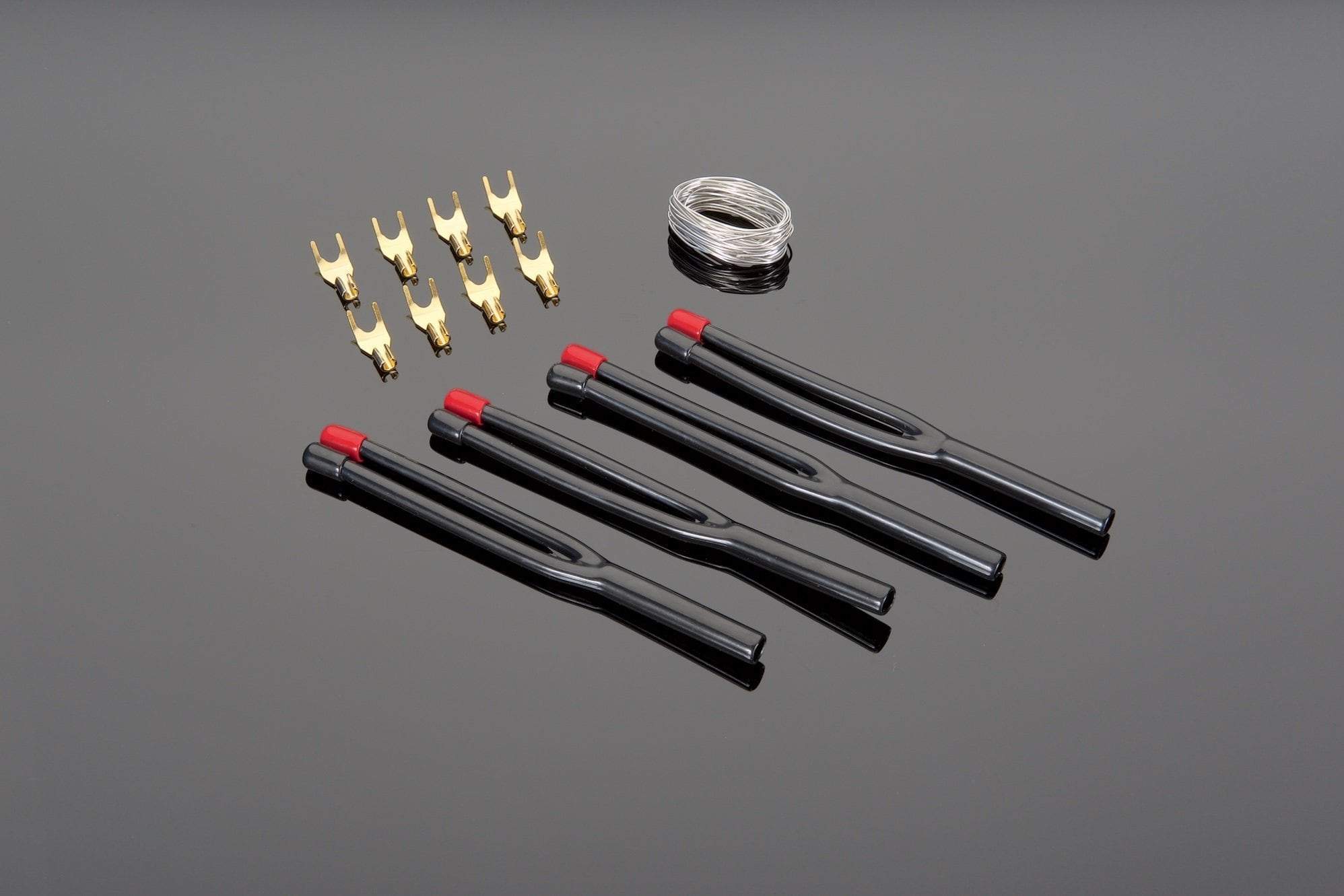 The image displays components from the Solder Termination Kits for High Performance 12-2 and 14-2 DIY Speaker Cable by Transparent Audio, including four black fuse pullers with red tips, a small coil of High Silver Content No Lead Solder, and eight gold-plated brass fork connectors, all placed on a reflective surface.