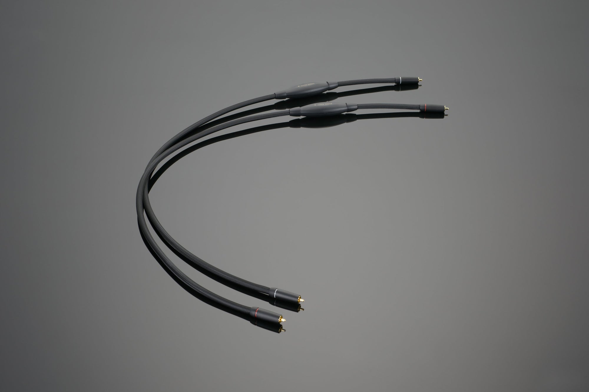 A close-up image of three Transparent Audio SUPER Phono Interconnect cables with gold-plated connectors, arranged in a curve on a plain, gray background.