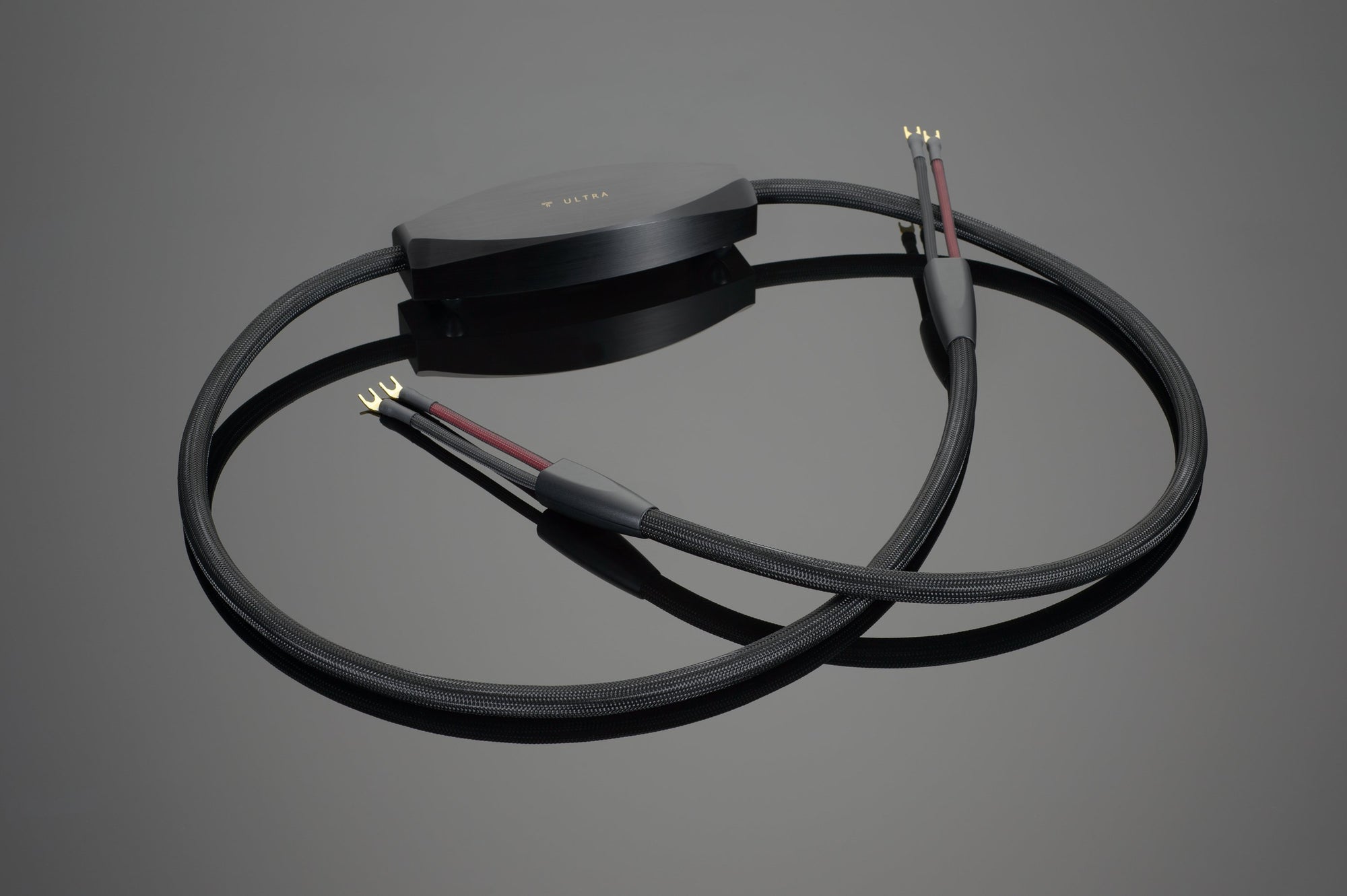 The Transparent Audio ULTRA Speaker Cable in black, featuring gold-plated connectors and a central black plastic housing, is displayed on a reflective surface.