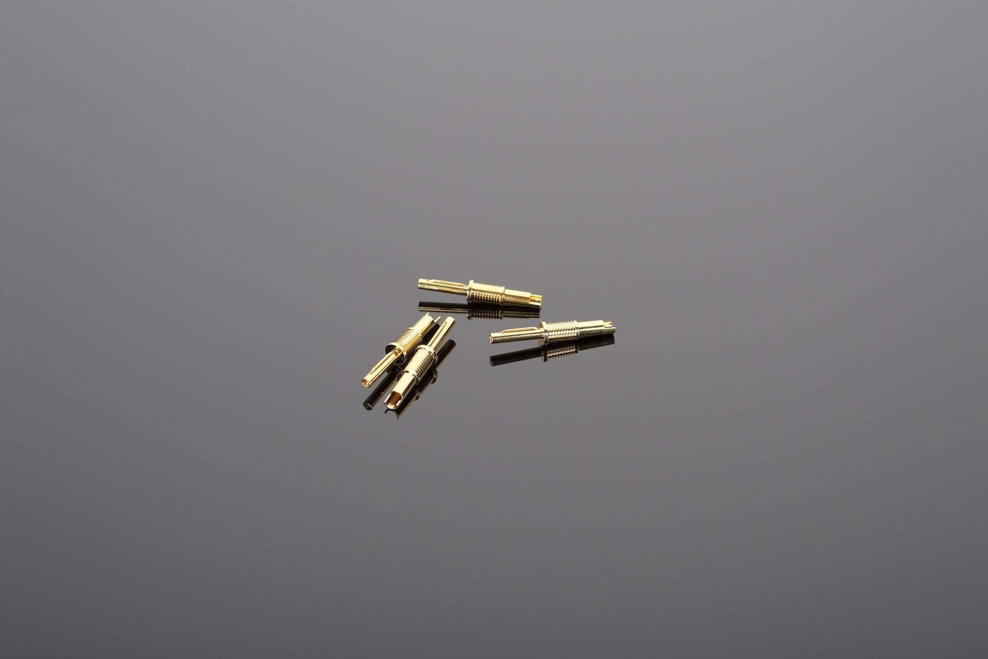 Several Universal Banana Connectors for soldering by Transparent Audio, featuring a small, gold-plated cylindrical design, are scattered on a plain, reflective surface.
