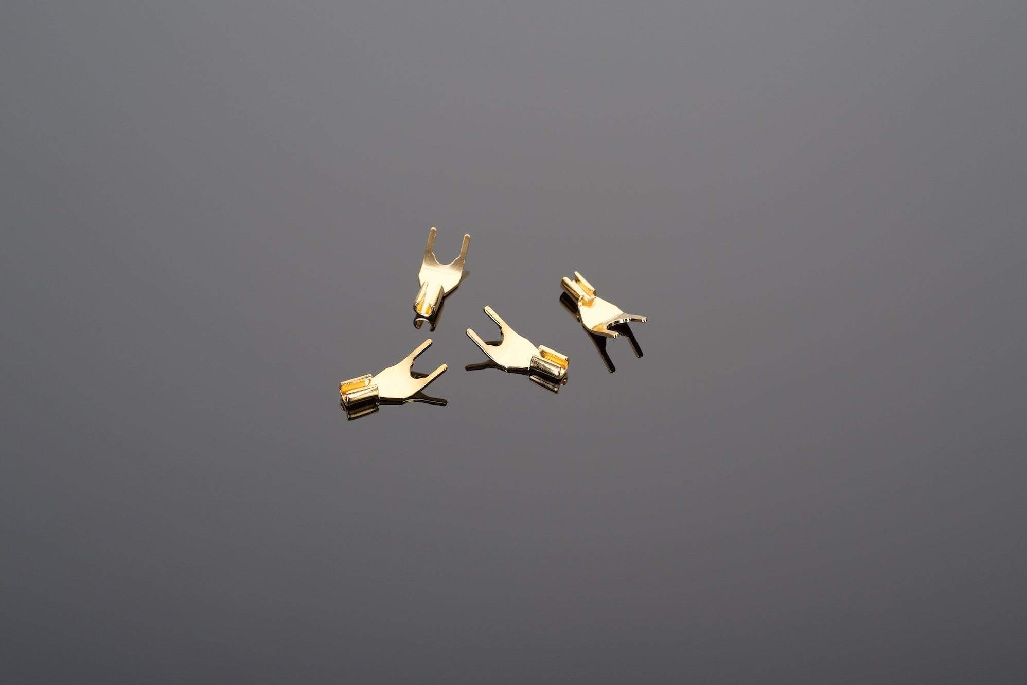 Four Universal Spade Connectors for soldering by Transparent Audio, small and gold-plated, are arranged on a smooth, reflective surface.